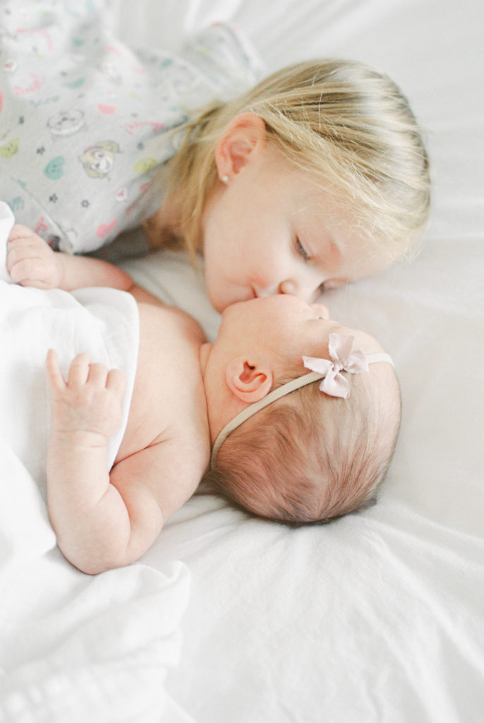 Two sisters laying on bed snuggling. Can you do your own newborn photoshoot? Yes, but it's not recommended.