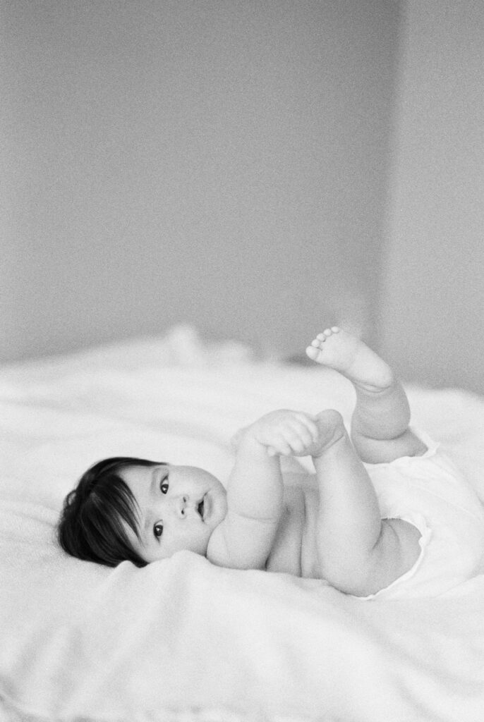 Six month milestone session near me. Baby laying down on bed holding toes. Captured on black and white film