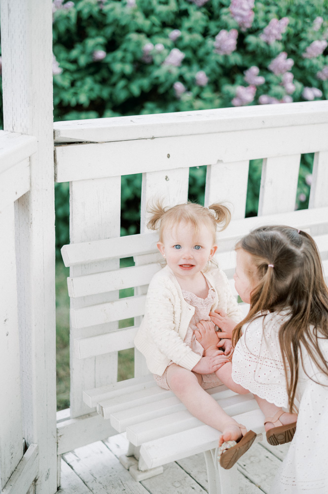 Two sisters snuggling up together in a white gazebo by a lilac bush