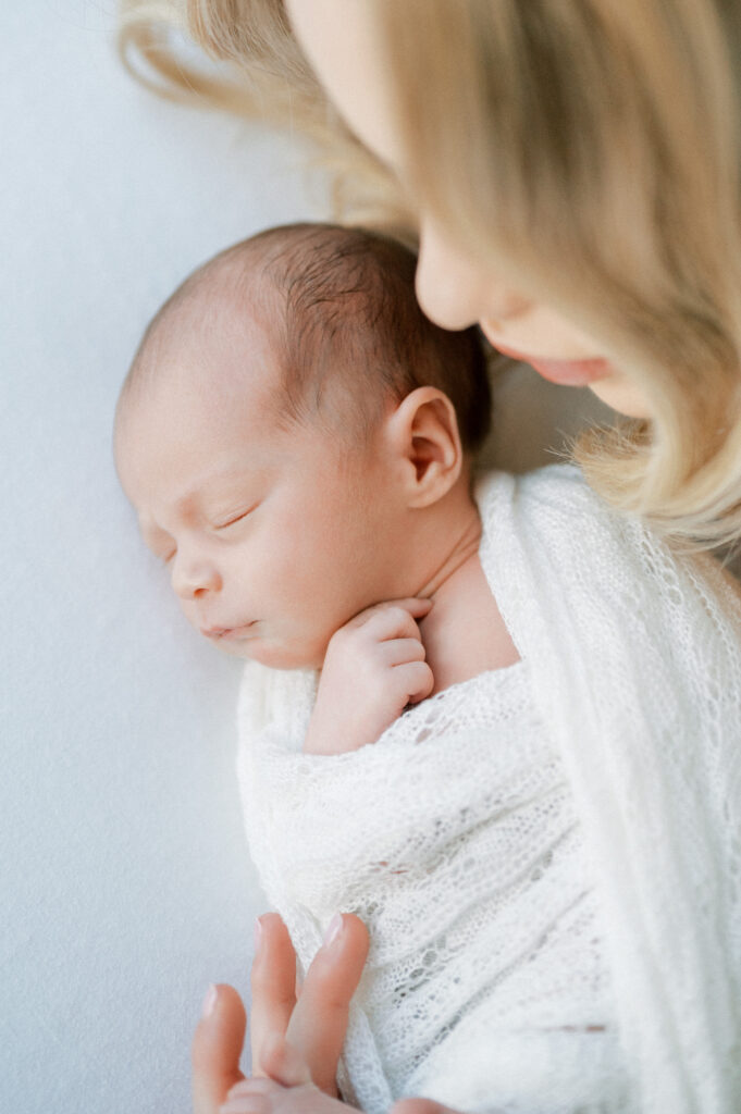 Baby wrapped in white blanket, snuggling close to mom during newborn photography session.